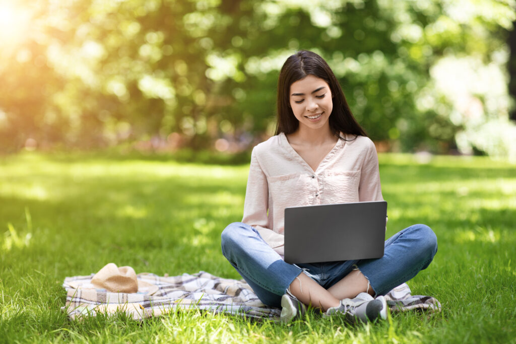 freelance concept. smiling asian girl working on laptop outdoors, sitting on lawn